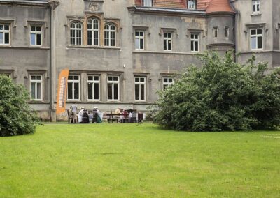 Pałac w Nakle Śląskim od frontu, na pierwszym planie trawnik. Przed pałacem ludzie w strojach z epoki siedzący przy stolikach. Po ich lewej stronie flaga z napisem Industriada.