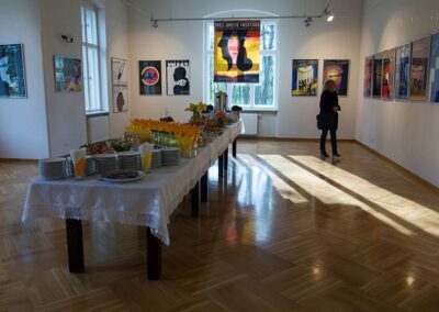 Sala wystawowa, w której eksponowane są kolorowe plakaty. Z prawej strony długowłosa blondynka przygląda się pracom. Na środku zastawiony stół cateringowy.