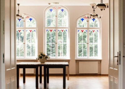 Otwarte drzwi do sali, w której na środku stoi duzy drewniany stół z obrusem i kompozycja z kwiatów. W tle trzy gotyckie okna z witrażami.