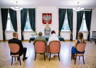 Cztery osoby siedzące tyłem na obitych, czerwona tapicerka krzesłach. Przed nimi stoi urzędzniczka stanu cywilnego. Z tyłu na scianie godło Polski.