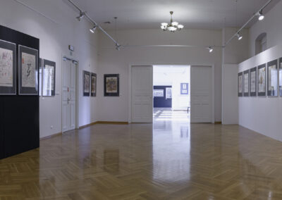 Duża sala wystawowa, po lewej stron tle białej ściany z wiszacymi grafikami. Po prawej stronie czarna scianka wystawowa z wiszącymi kolorowymi grafikami. Na wprost duże rozsuwane drzwi za którymi widać salę rozetową.